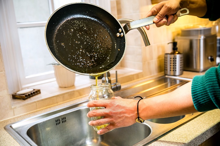 A person in a green jumper pours oil from a frying pan into a glass jar.