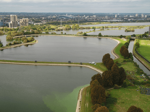 A birds eye view across two reservioirs with green trees and grass on the right of the picture and tall buildings in the background.