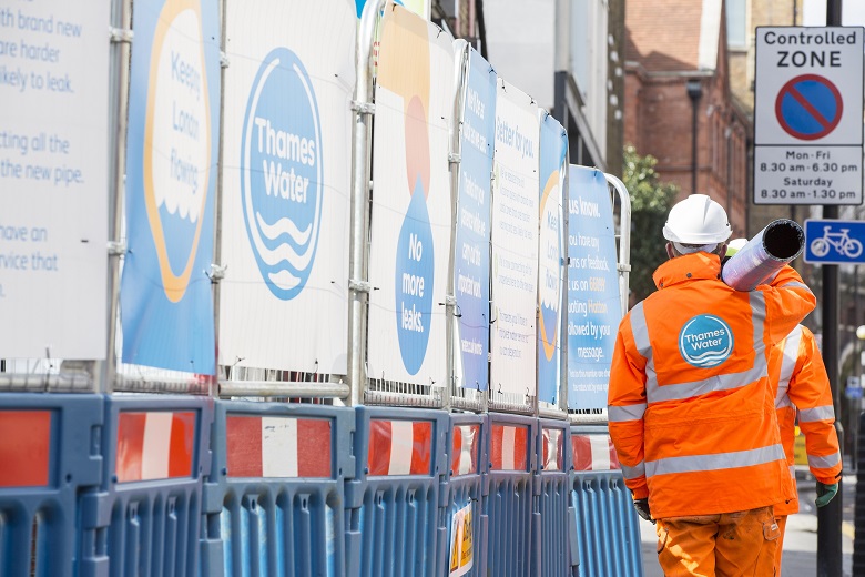 Two engineers wearing orange overalls carry a pipe in front of a Thames Water sign.