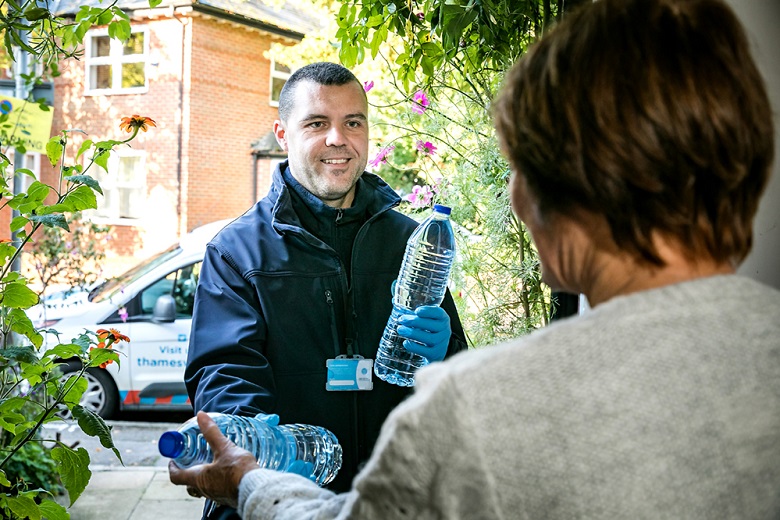 A Thames Water employee dressed in blue hands bottled water to an older lady in grey jumper