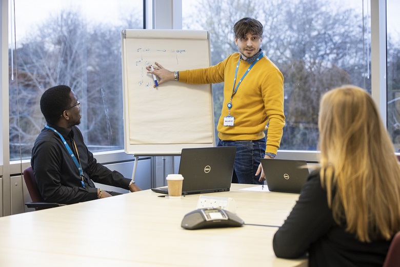 A man in a yellow jumpers speak to two colleagues.