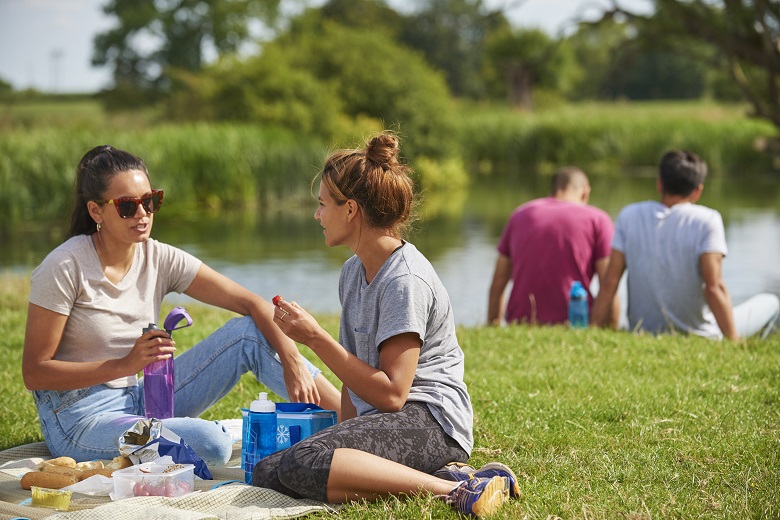Two friends sit on a picnic blanket next to a river