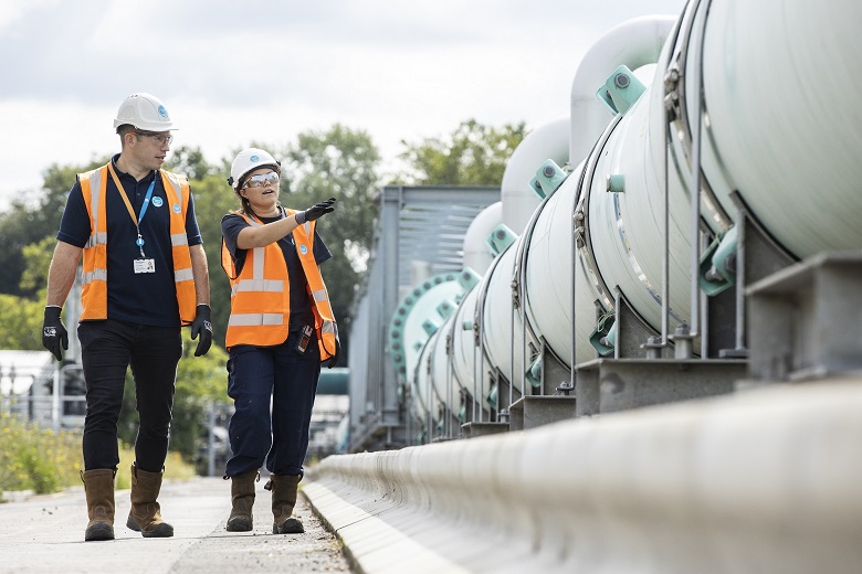 A man and a woman wearing orange high-vis walk past a grey air main.