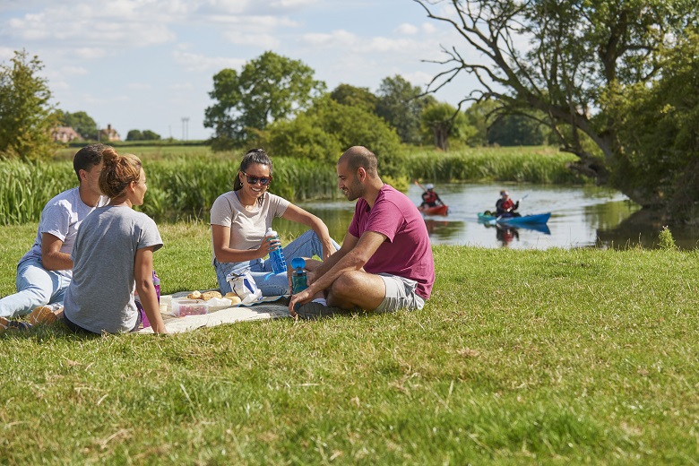Four people sit with a picnic next to a river which has two kayakers paddling on it
