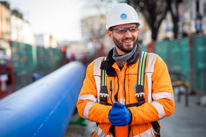 Man in orange high vis smiles and stands in front of a large blue pipe.