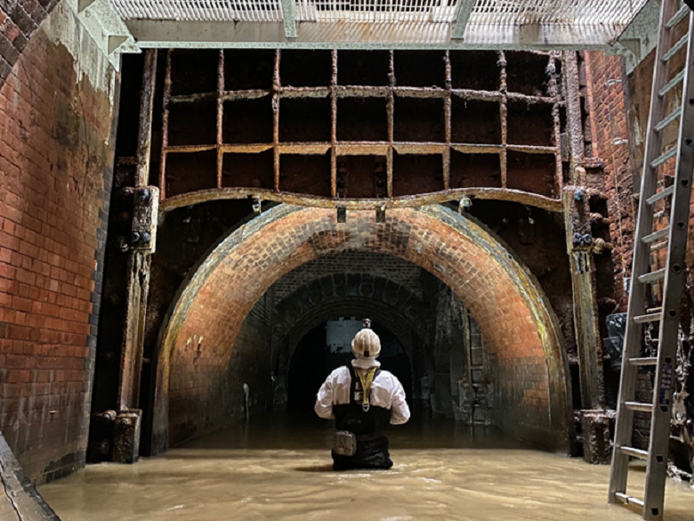 A man in black and white protective clothing stands waist deep in the middle of a sewer