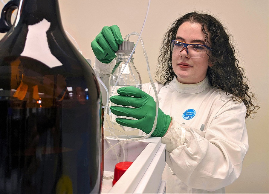 A woman with dark hair working in a lab wearing a lab coat and protective glasses