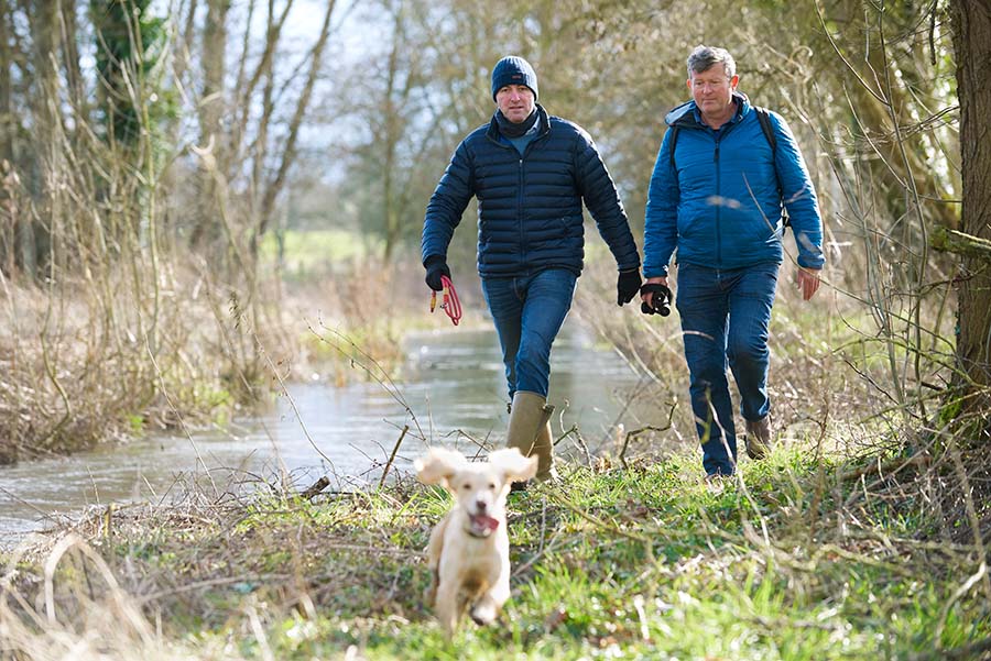 Two people walk alongside river carrying binoculars while their dog runs in front