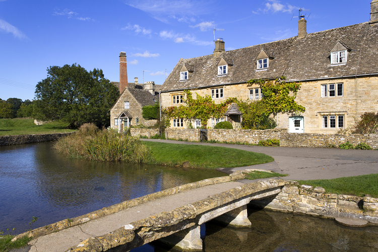 A row of stone cottages with a river and a small stone bridge in the foreground. 