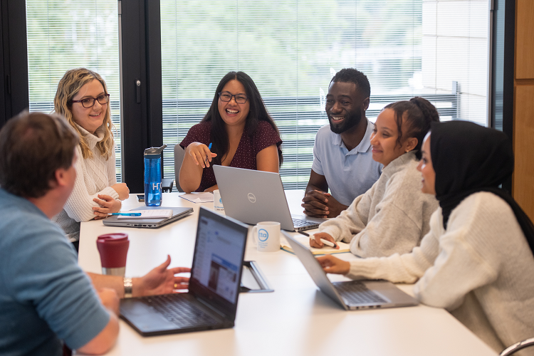 A group of people sitting at a table having a working meeting with laptops open in front of a window.