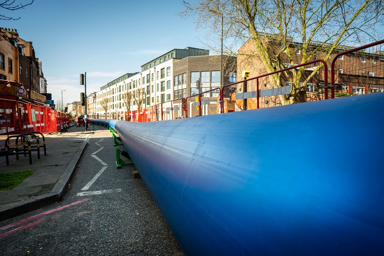 A road with tall buildings in the distance and a large blue plastic pipe in the front.