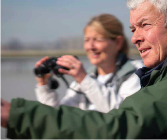 A man in a green jacket pointing beyond the shot with a woman in a white jacket with a pair of binoculars.