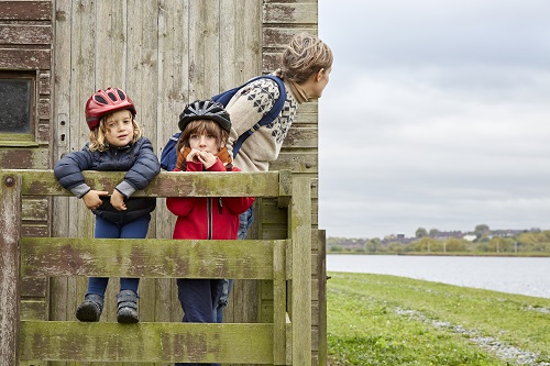 Two children looking at the camera wearing cycle helmets on a fence while an adult woman looks around the side of a wooden building.
