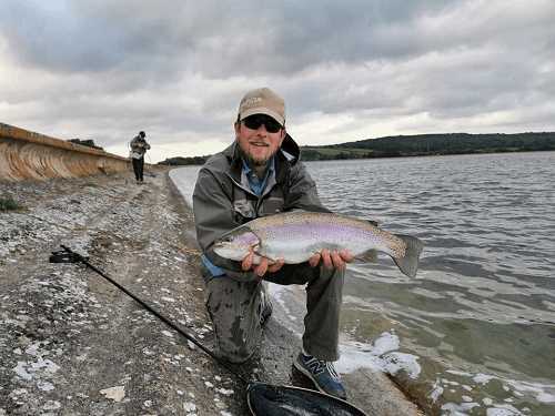 A man wearing a cap and sunglasses holding a large fish next to water with a fishing rod on the sloping bank.