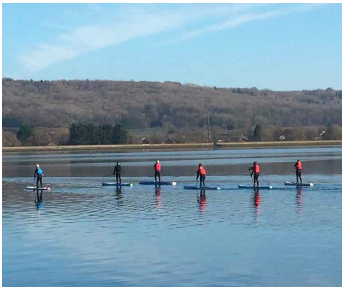 A group of people on paddle boards on a body of water with trees on the bank in the distance under a blue sky.