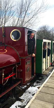 A small red train engine with green and cream coloured carriages behind and the station platform with a man in the drivers cab.