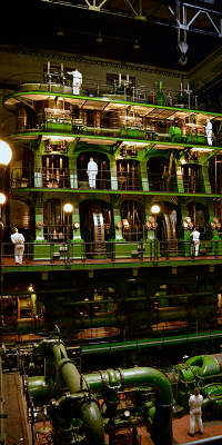 A large mechanism of green pipes at floor level with three further levels of large wheels and pistons with some people standing on each level.