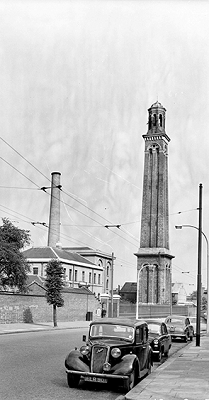 A black and white photo of a large brick tower with old cars parked on the road in front and a chimney in the distance.