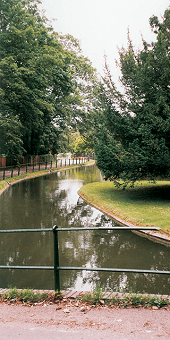 A channel of water into the distance surrounded by green trees with a footpath along the left side and a green metal fence in front. 