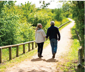 A man and woman hold hands walking along a footpath between woodland bushes and grassland in bright sunshine.
