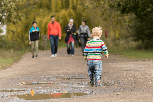 A child with blonde hair in a stripy jumper and wellies walks past a puddle in the path with other people in the distance. 