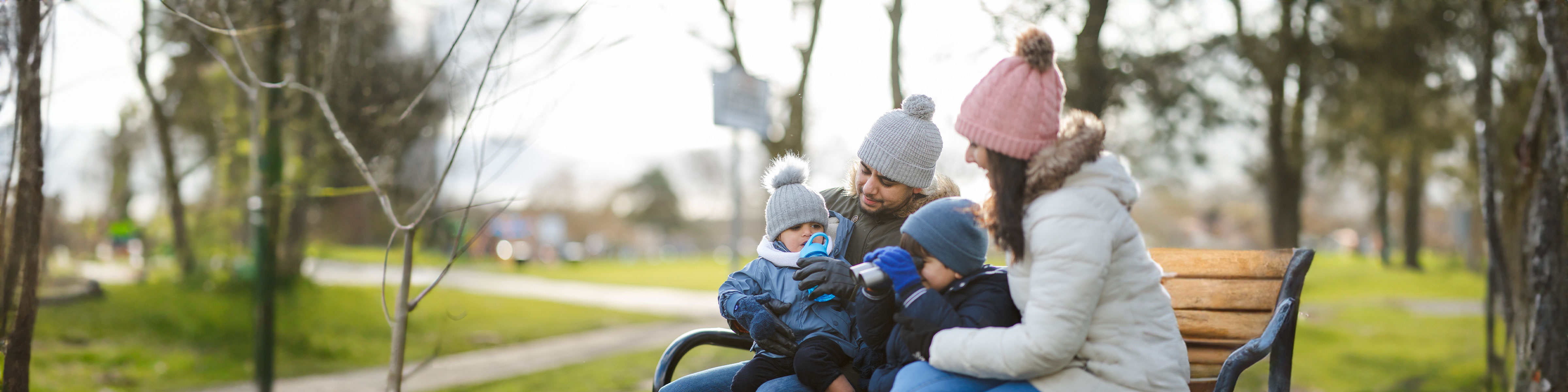 A young family sit on a bench in a park with winter hats on