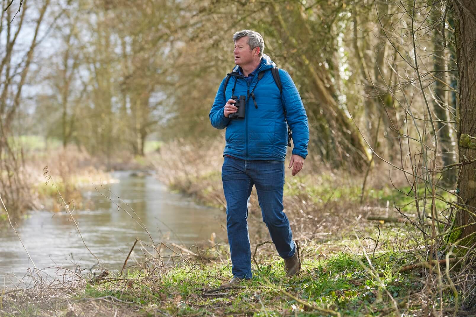 A man walks alongside a river with binoculars
