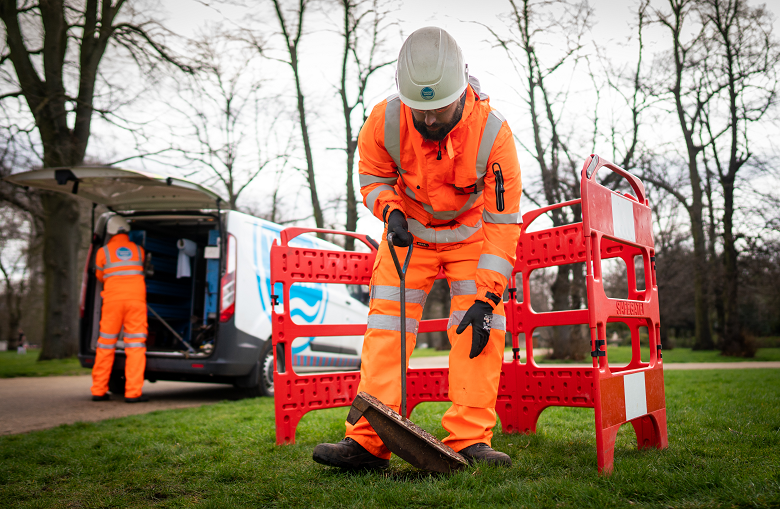 A person in orange high-visibility clothing and a white hard hat using a tool to lift a drain cover surrounded by orange fencing.  