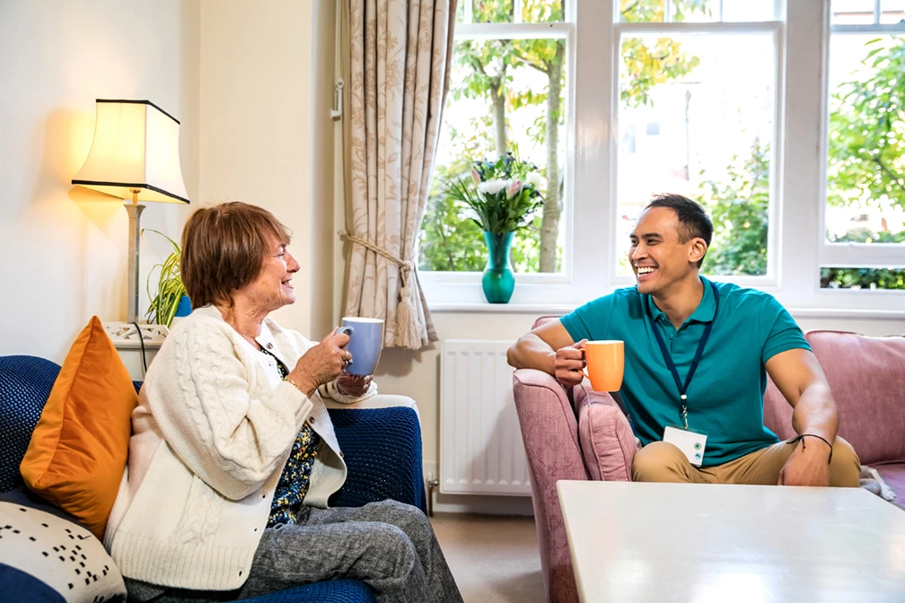 An elderly woman and their carer, a man wearing a green top, enjoy a cup of tea together sitting on the sofa